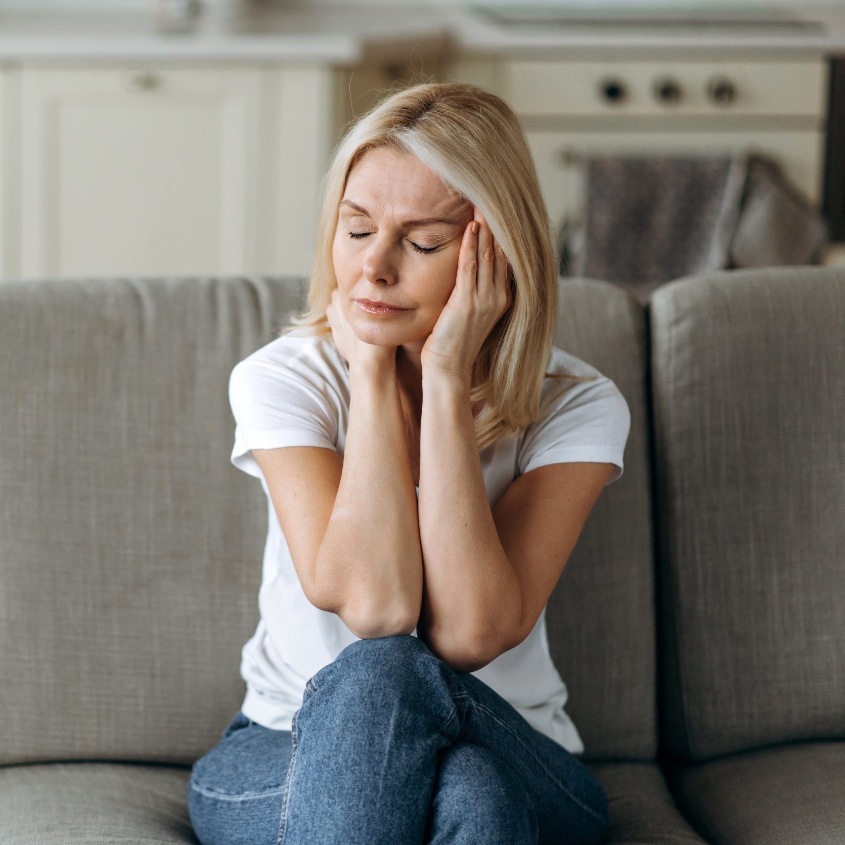 Headache, depressed or stress. Middle aged blonde woman sits on sofa at living room holding her head with her hands, feels unhappy because of headache, stress, depressed or bad news, she need rest and support